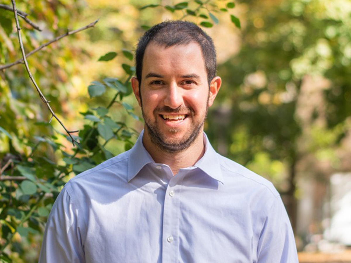  Physics professor Aidan Brown stands outside looking at the camera on a sunny day in front of a green hedge